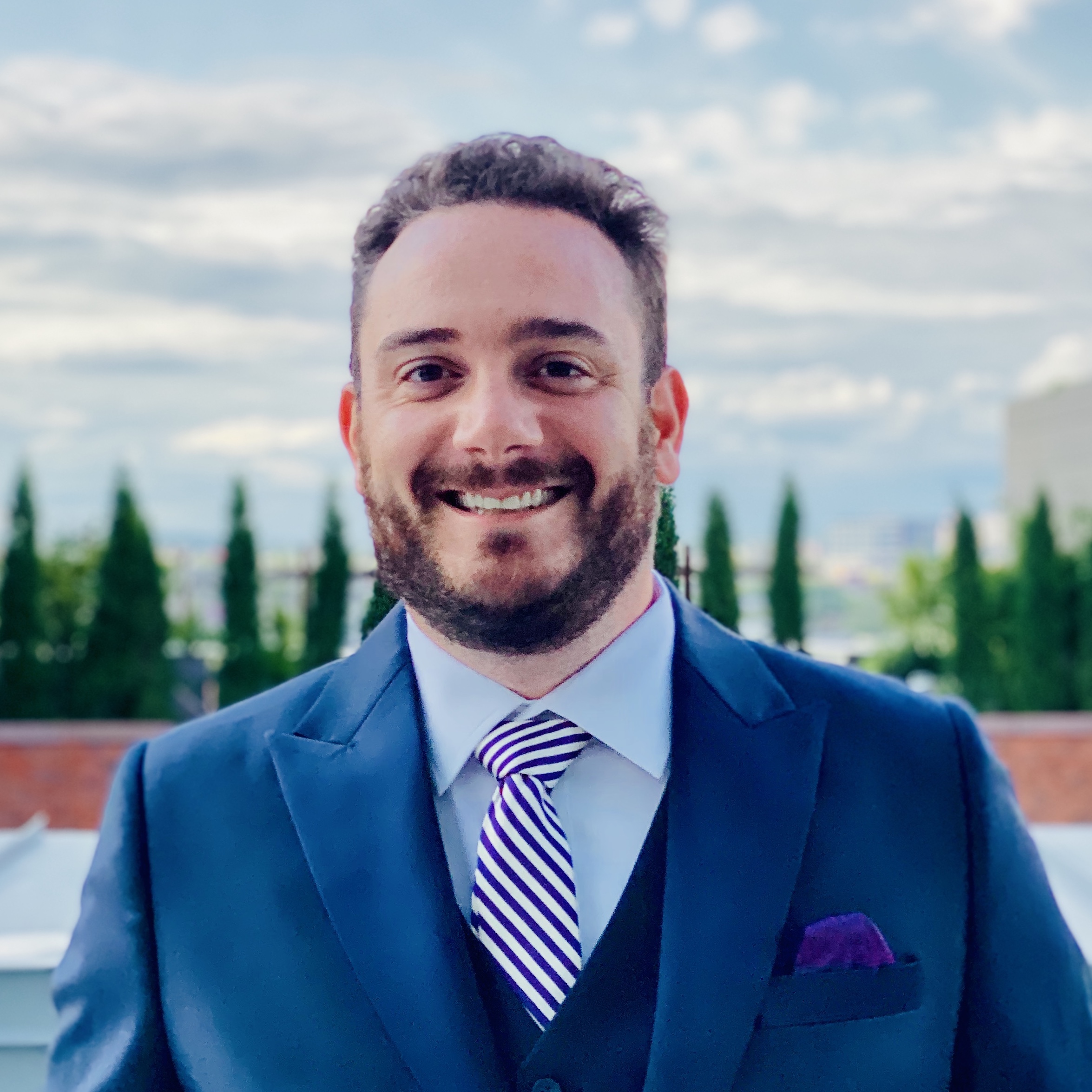 Robert Calise smiling outdoors in a navy blue suit with purple striped tie and pocket square, with trees and blue sky in the background.